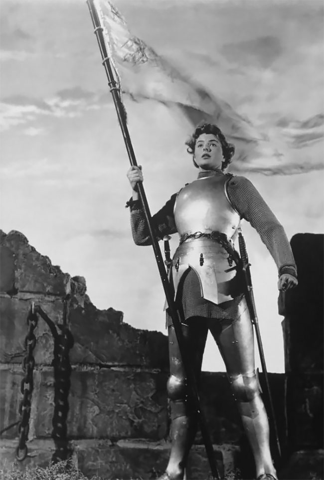 Black and white photo of actress Ingrid Bergman dressed in knight's armour and holding a banner. Sky and castle wall in background.