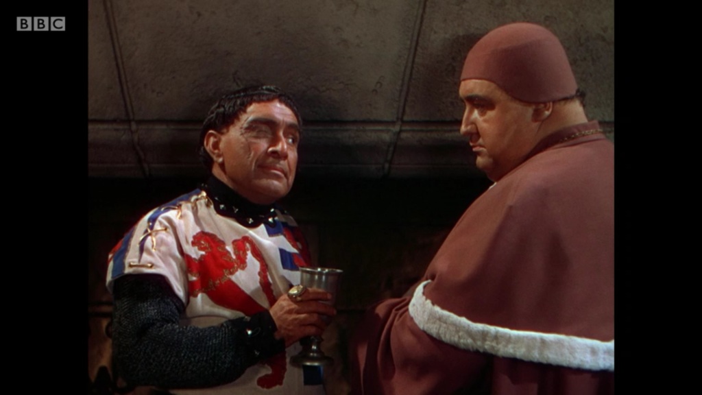 Film image of two medieval men looking at each other: one has dark hair, a tunic with a red dragon on it, and a large scar onhis face, while the other is a fat man in a rust-coloured bishop's costume.